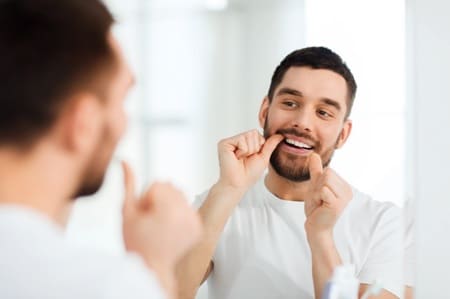 Man with facial hair leaning over skink to floss teeth in the mirror
