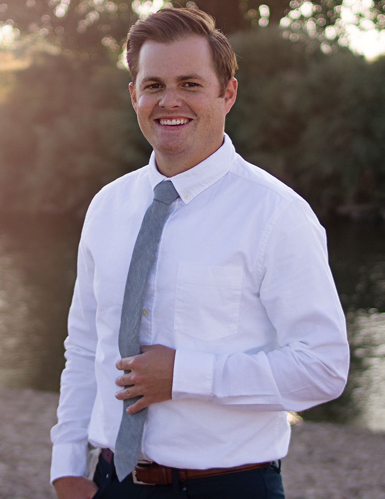 headshot of man in white collared shirt and tie, holding his tie in one hand and smiling, with nature background